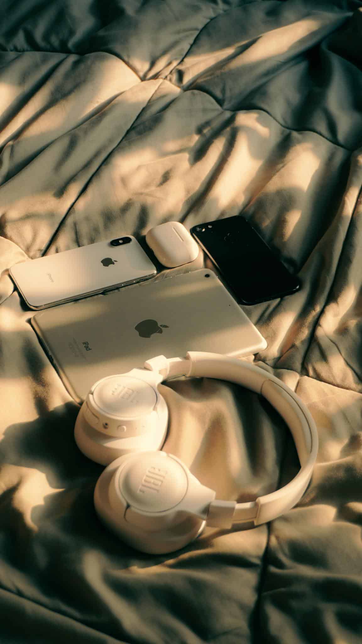 A collection of Apple devices including MacBook, iPad, and iPhone on a bed with sunlight.